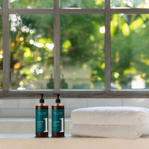 Two amber pump bottles of TRUE MOUTH cleansers sit on a bathroom counter next to a stack of white towels, with a blurred window and green foliage in the background.