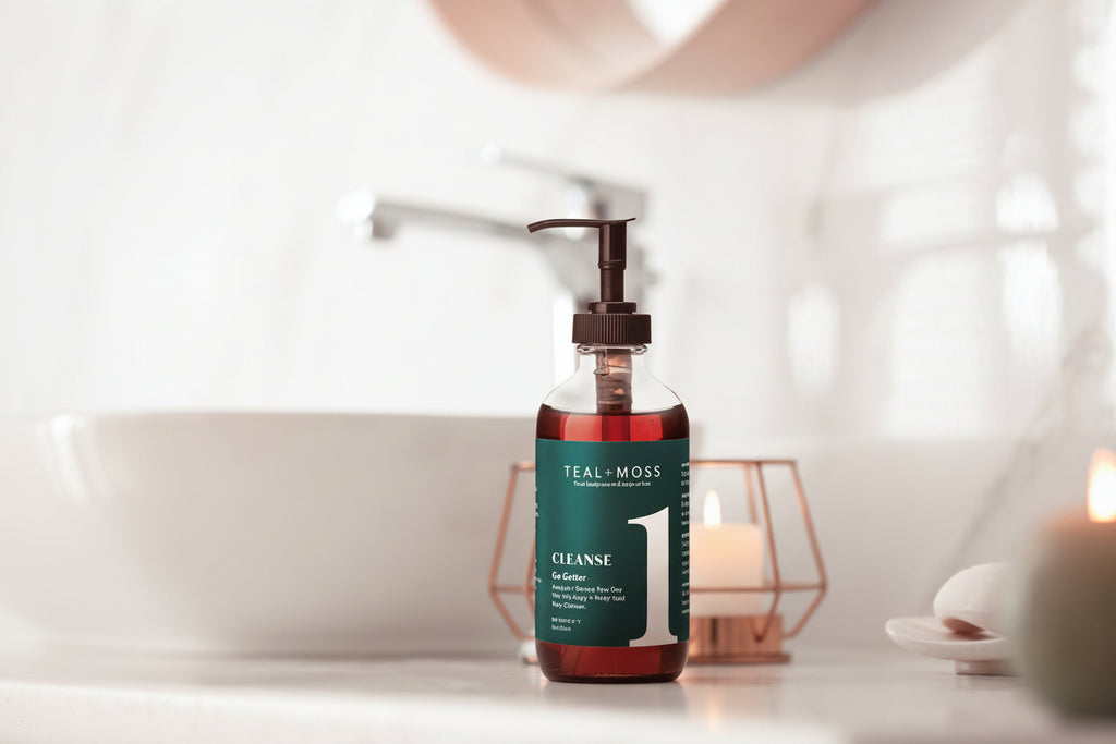 Two Teal-Moss skincare bottles on a bathroom counter: a larger amber bottle with a teal label and pump labeled 'CLEANSE 1' with a blurred sink and faucet in the background.
