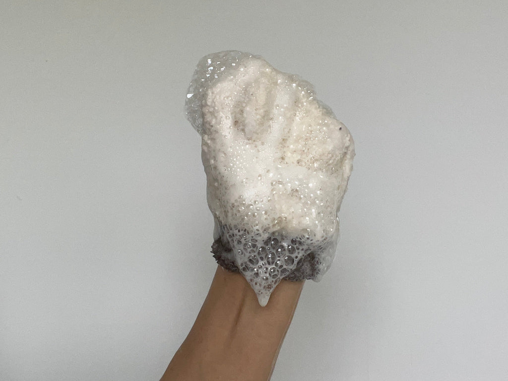 A soap foam covered loofah to show product consistency of Teal and Moss Cleaners, raised against a plain light gray wall.