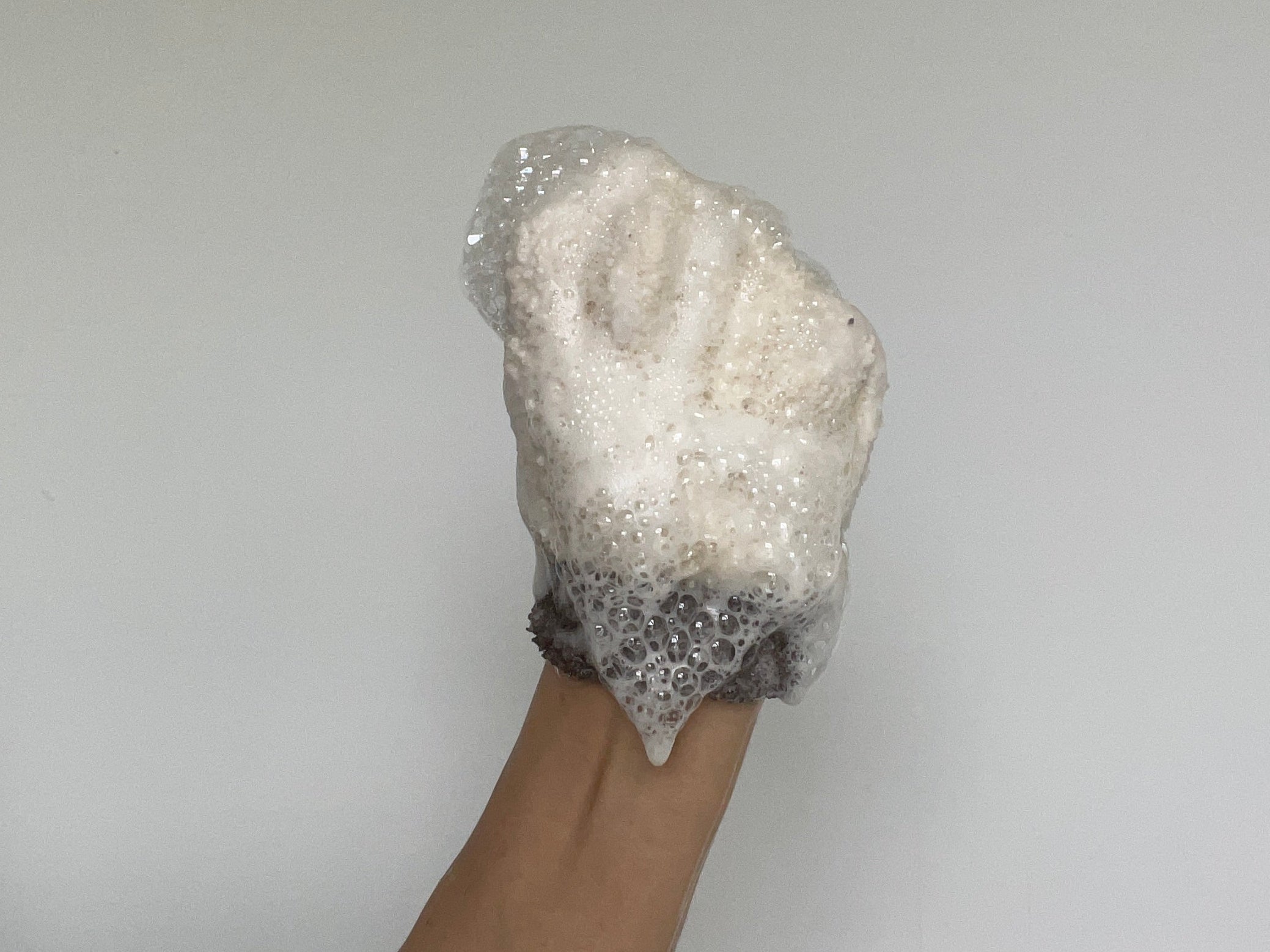 A soap foam covered loofah to show product consistency of Teal and Moss Cleaners, raised against a plain light gray wall.
